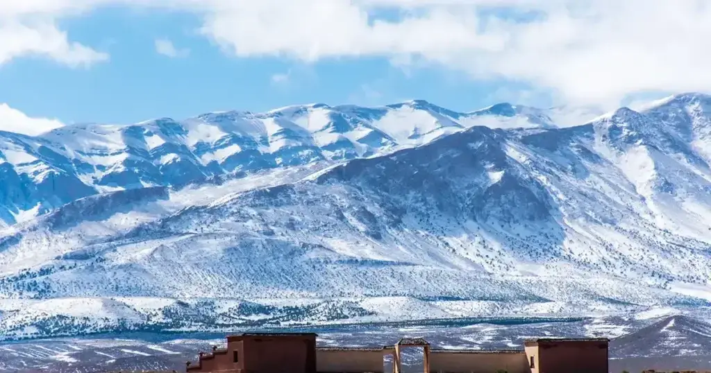 Snow-capped peaks of the High Atlas Mountains rising above a traditional earthen kasbah building on the open plain below, Morocco