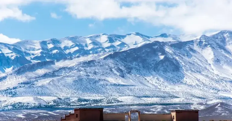 Snow-capped peaks of the High Atlas Mountains rising above a traditional earthen kasbah building on the open plain below, Morocco