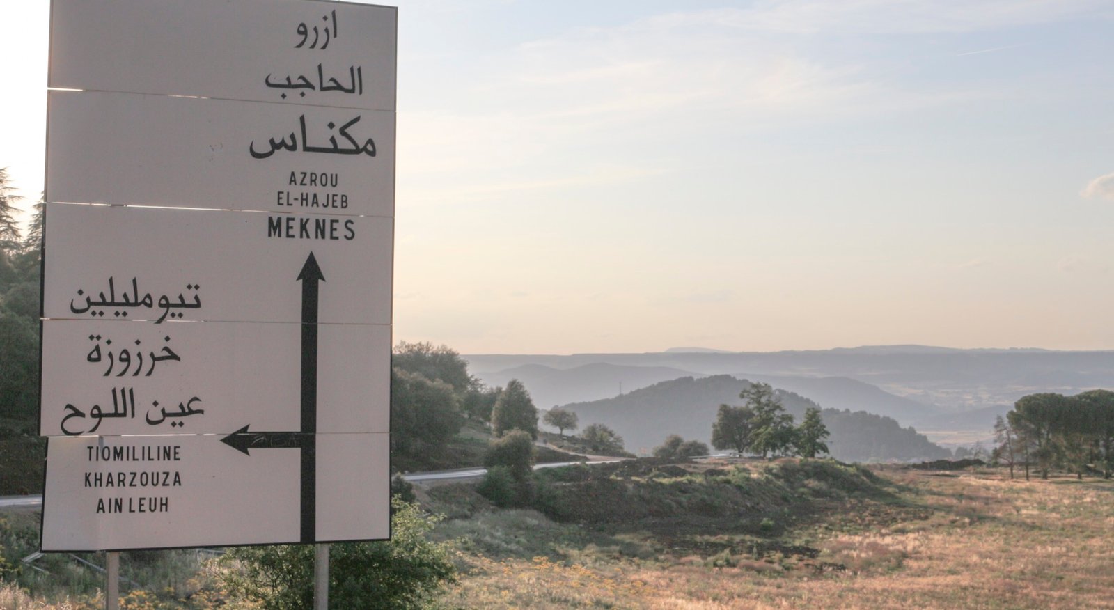 A bilingual Arabic and French directional road sign at a rural intersection in Morocco, showing distances to nearby towns